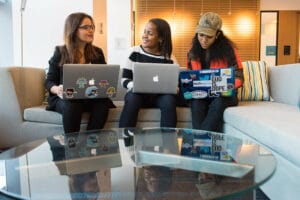 Tre donne siedono una a fianco all'altra sul sofa con il pc sulle ginocchia. Gli sticker sulle cover dei loro pc rivelano che probabilmente lavorano nel tech, ma le tre non sembrano arrabbiate anzi, sorridono.